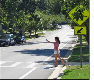 Photo shows a white painted crosswalk across a two-lane street with bike lanes on both sides and parking on the far side.  A post in the grass near the crosswalk has a diamond-shaped yellow sign with a sillouette of a pedestrian walking, and a square yellow sign below that has an arrow pointing down to the crosswalk.  Joumania is standing with one foot in the street and the other foot at the curb, wearing a checkered red and white blouse and white shorts.  Her arm is straight out holding a white cane pointing up to the sky and slightly forward.
